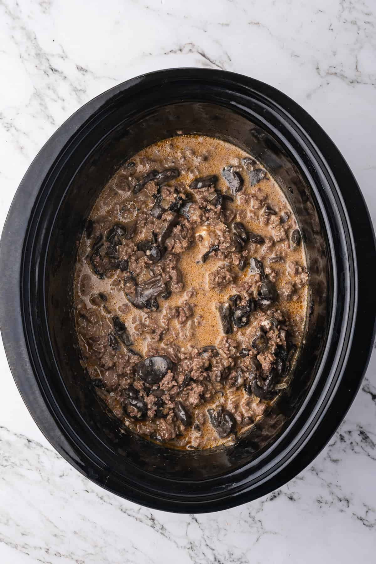 Ground beef, black olives, and a savory sauce mixture simmer together in a black slow cooker, perfect for creating an easy Slow Cooker Beef Stroganoff on a marble countertop.