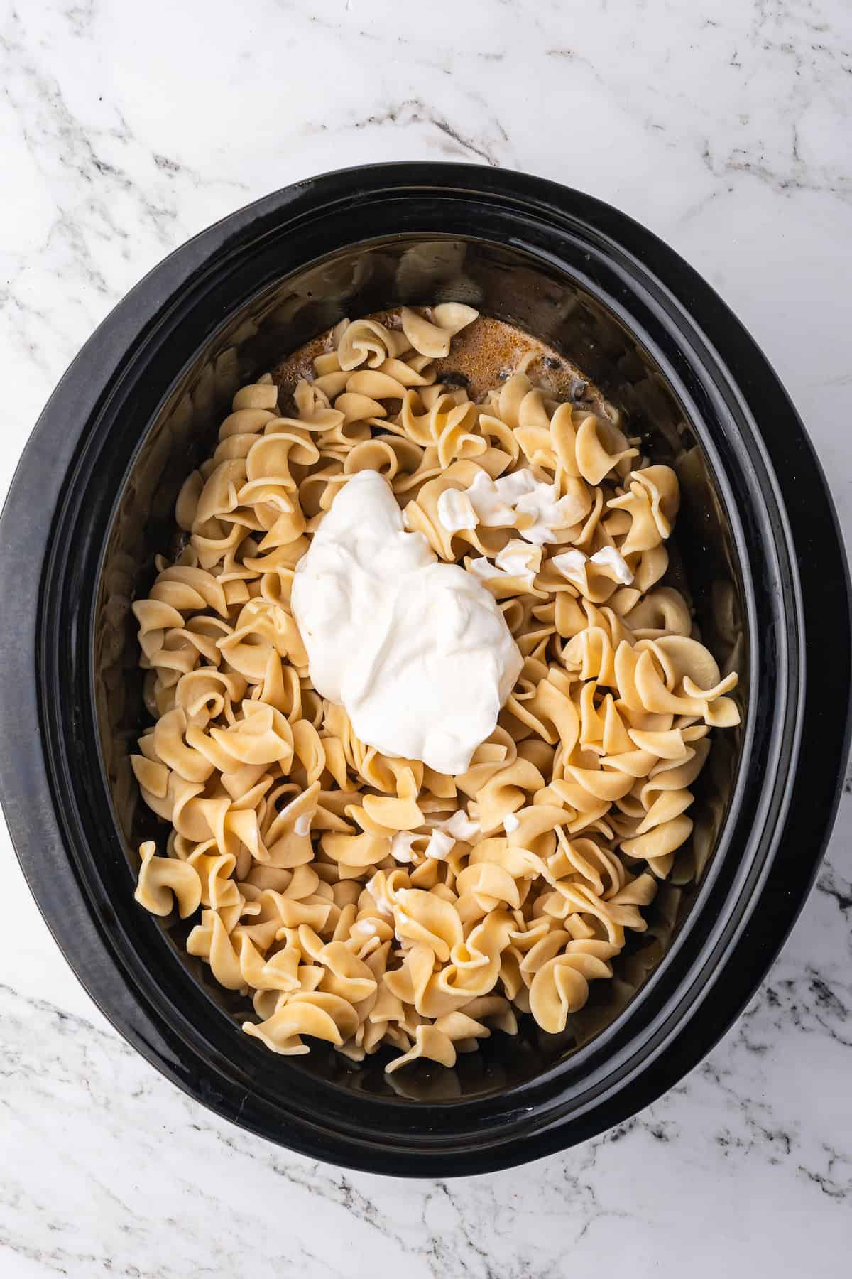 Uncooked egg noodles and a dollop of sour cream sit in a black slow cooker on a marble countertop, ready to become delicious Slow Cooker Beef Stroganoff.