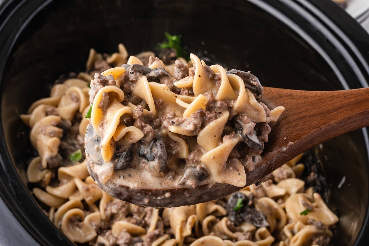 A wooden spoon holds a portion of creamy Slow Cooker Beef Stroganoff with egg noodles, mushrooms, and ground beef, above a slow cooker brimming with the savory dish.