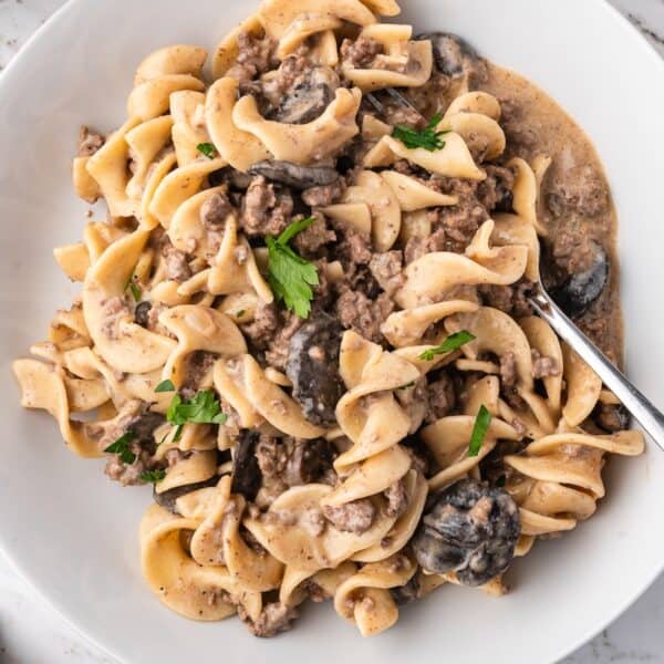 A plate of Slow Cooker Beef Stroganoff with egg noodles, ground beef, mushrooms, creamy sauce, and parsley garnish is served with a fork.