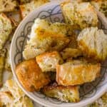 A bowl filled with seasoned sourdough croutons is surrounded by more croutons on a patterned surface.