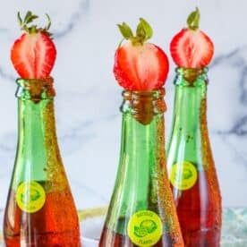 Three green glass bottles with brown liquid are topped with halved strawberries. Their rims are coated with chili powder and seasoning, making them perfect for a refreshing strawberry margarita topo chico twist.