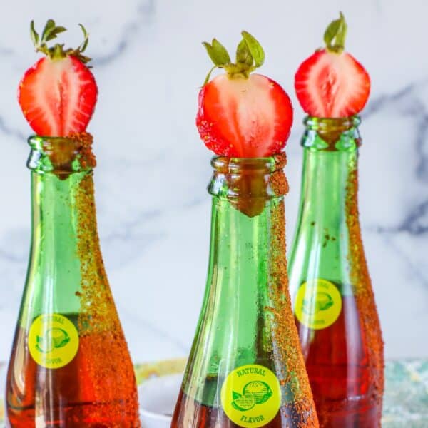 Three green glass bottles with brown liquid are topped with halved strawberries. Their rims are coated with chili powder and seasoning, making them perfect for a refreshing strawberry margarita topo chico twist.