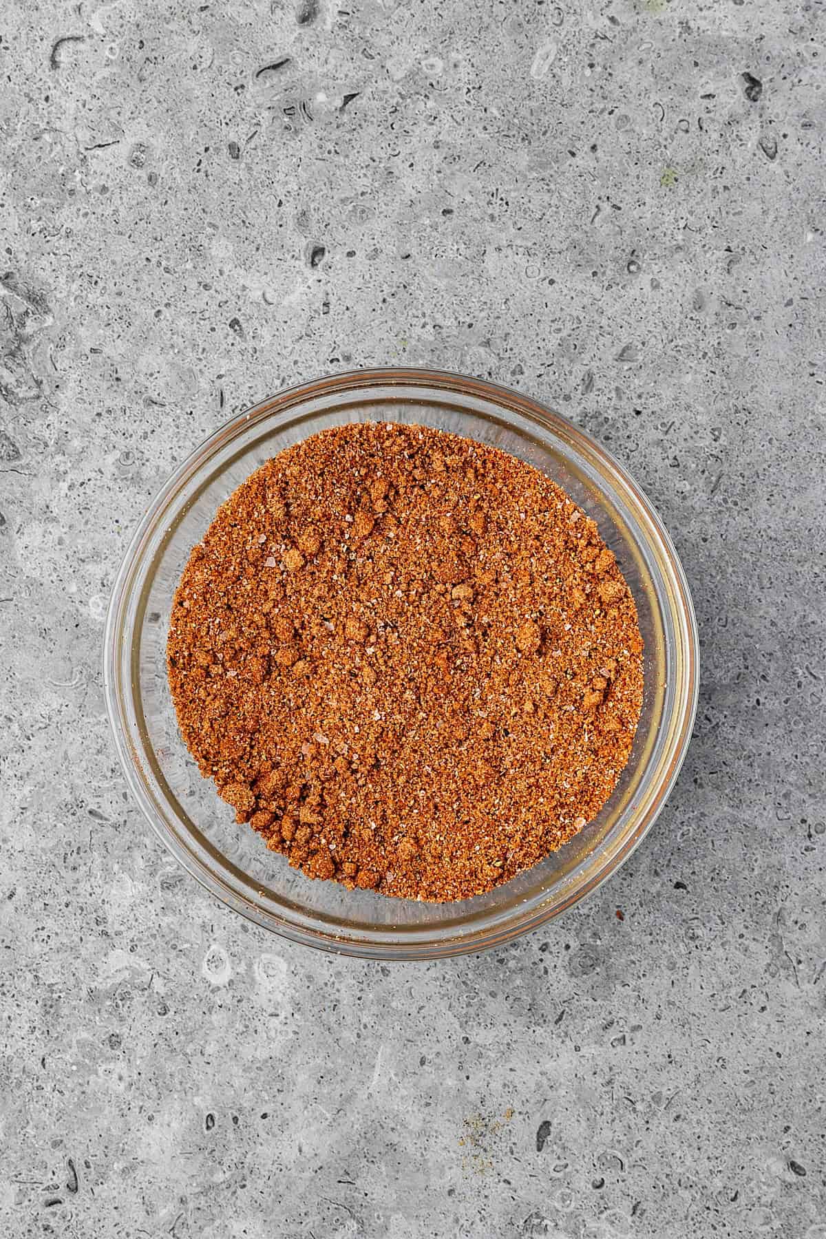 A clear glass bowl filled with a reddish-brown smoked BBQ spice mixture sits on a gray, textured stone surface.