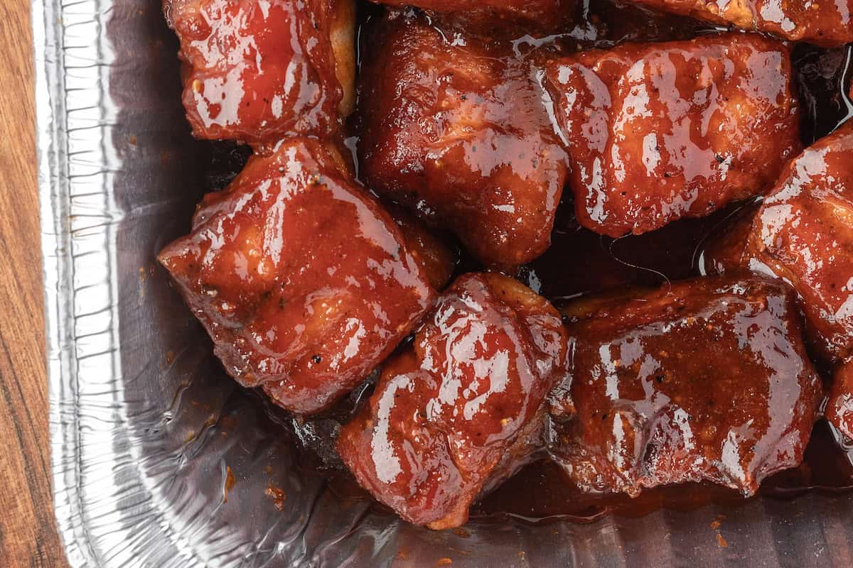 Close-up of glazed, saucy BBQ Pork burnt ends in a disposable aluminum tray on a wooden surface—these mouthwatering pork belly bites are smoked to perfection.