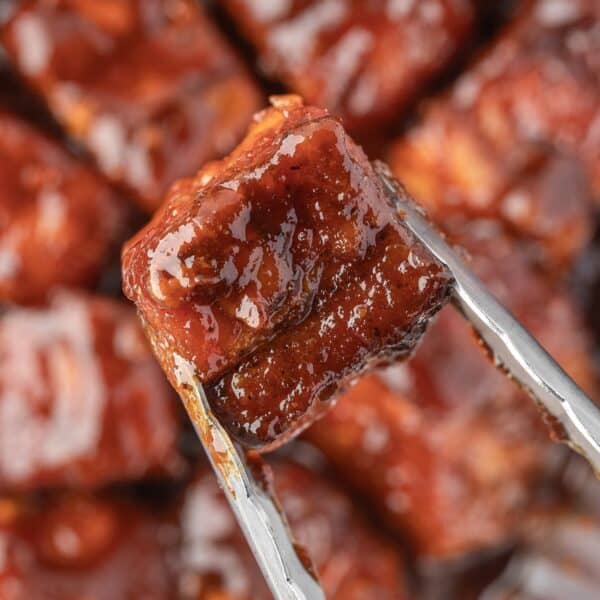 Close-up of metal tongs holding a glazed, smoked pork belly cube with more tender pork belly bites visible in the blurred background.