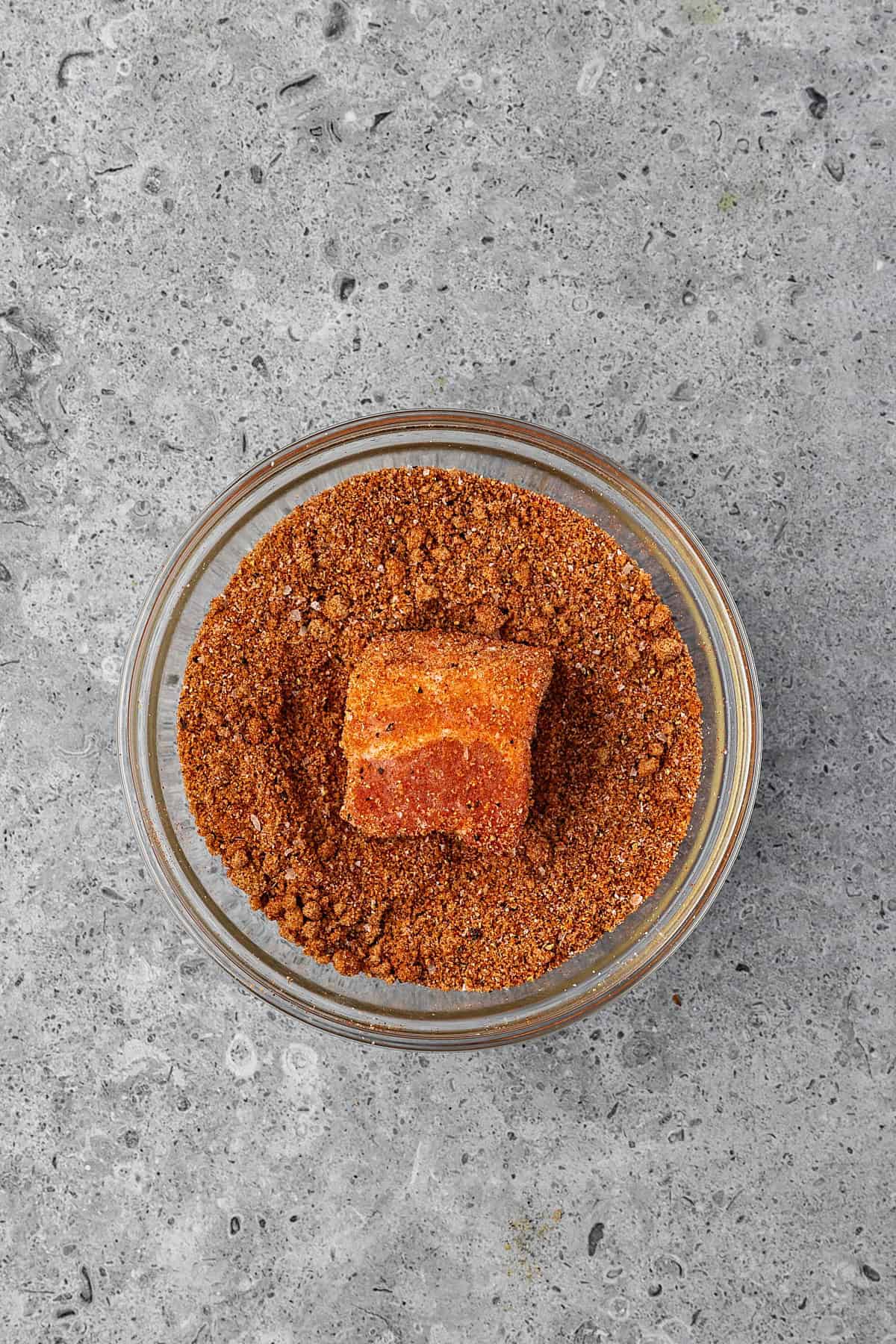 A piece of BBQ Pork Belly coated in a red-brown spice rub sits in a small glass bowl filled with more spice mix, placed on a gray textured surface.