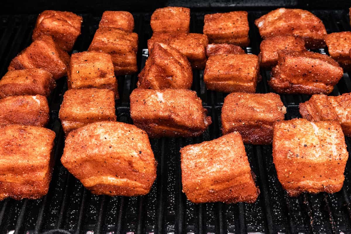 Chunks of seasoned Pork Belly Bites are arranged on a black grill, cooking with a visible outer spice rub, perfect for smoked BBQ flavor.