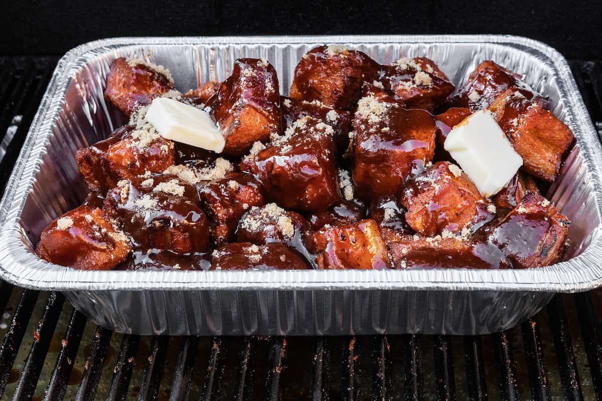A foil tray filled with BBQ pork belly bites covered in sauce, topped with pats of butter and sprinkled with brown sugar, placed on a grill.
