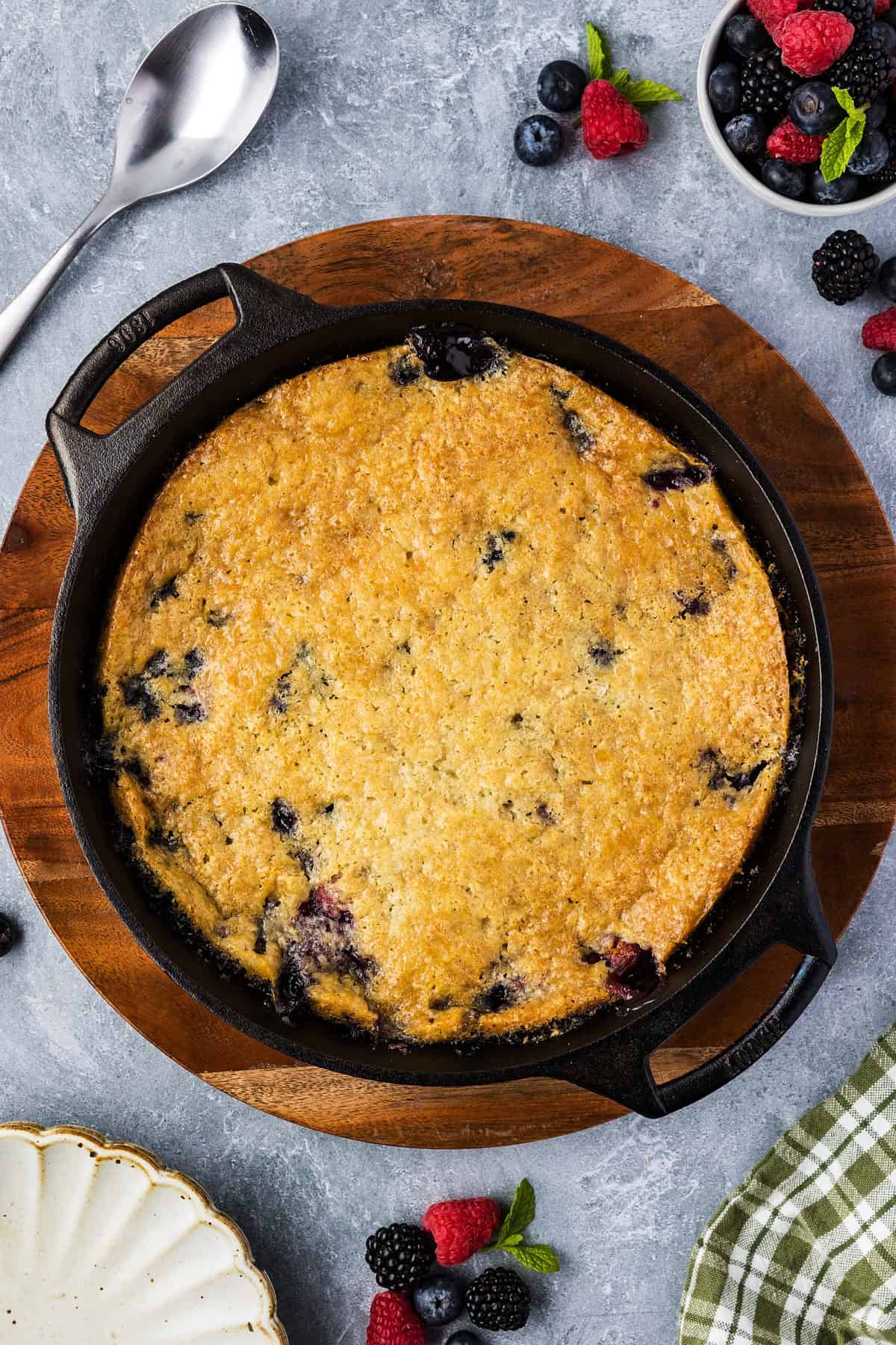 A freshly baked Smoked Berry Cobbler in a cast iron skillet sits on a wooden board, surrounded by fresh berries, a spoon, and a green checkered napkin.