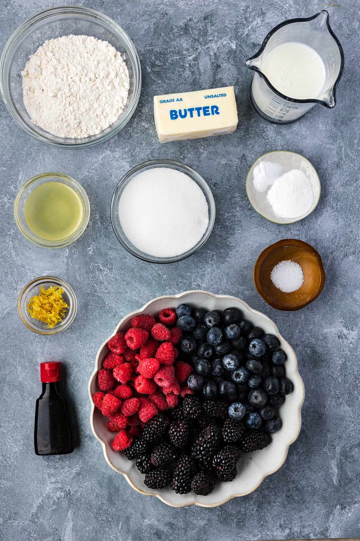 Various baking ingredients are arranged on a gray surface, ready to create a delicious Smoked Berry Cobbler—flour, butter, milk, sugar, blackberries, raspberries, blueberries, vanilla, lemon zest, oil, baking powder, and salt.