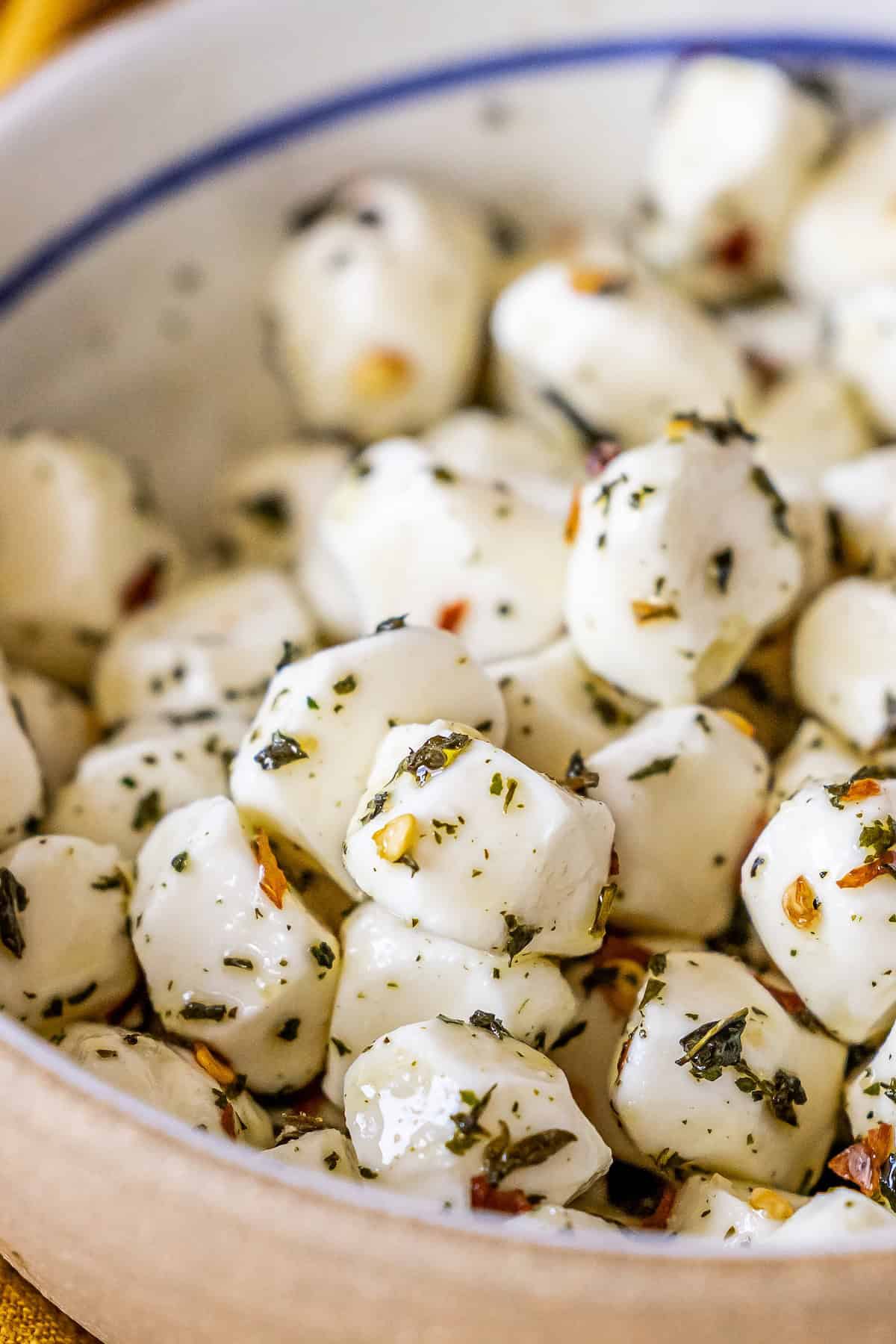 A close-up of marinated mozzarella balls coated with herbs and spices in a ceramic bowl.