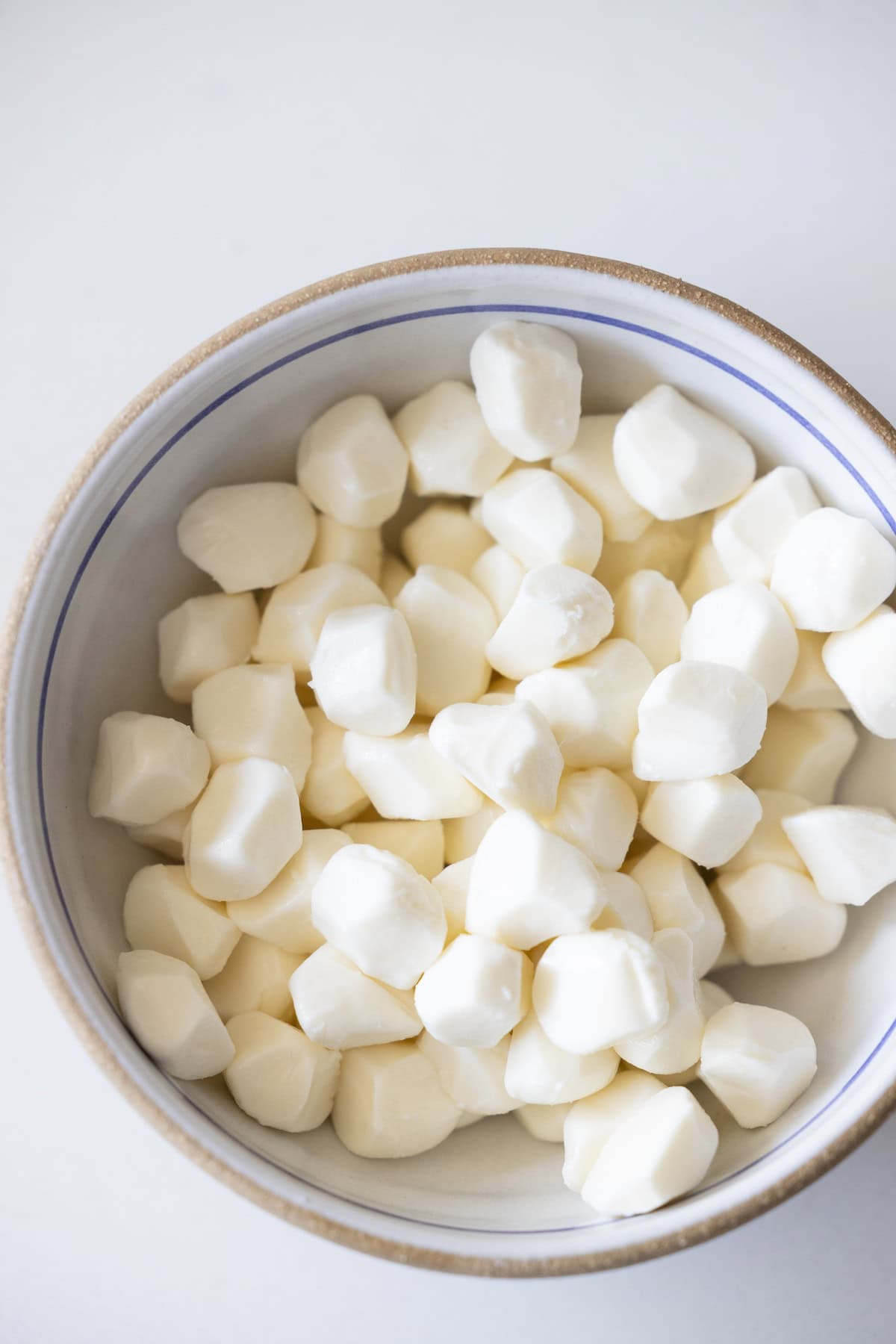 A bowl filled with small, white marinated mozzarella cheese balls on a white surface.