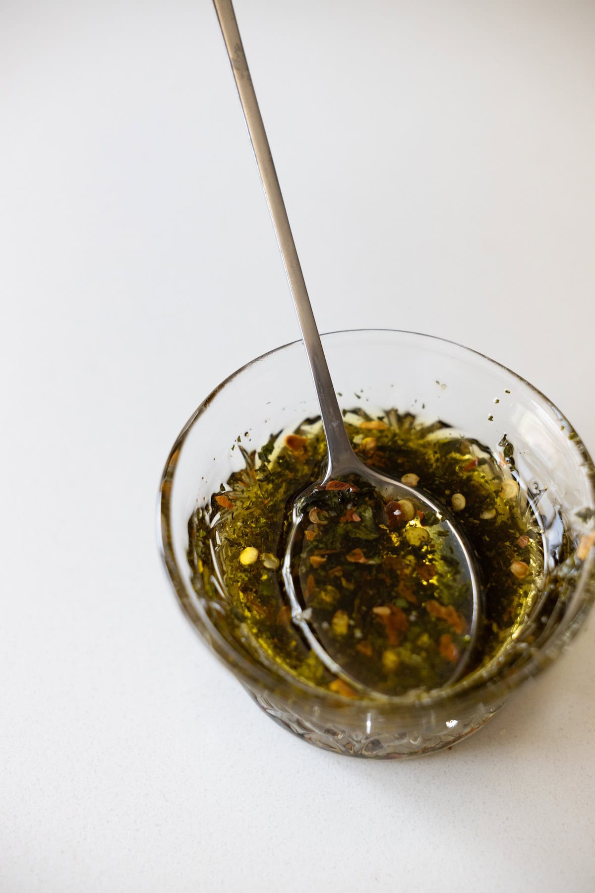 A metal spoon rests in a glass bowl filled with olive oil, dried herbs, and red pepper flakes—perfect for creating a delicious Marinated Mozzarella recipe on a white surface.