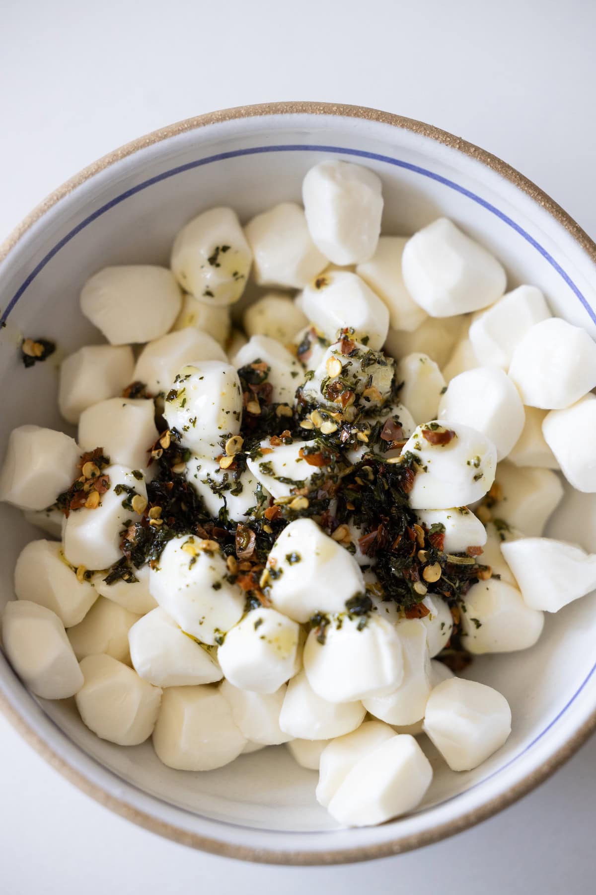 A bowl of marinated mozzarella balls topped with dried herbs and red pepper flakes on a white surface—a simple yet flavorful mozzarella recipe.