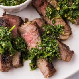 Sliced grilled steak with chimichurri, topped with vibrant green sauce and served on a white plate.