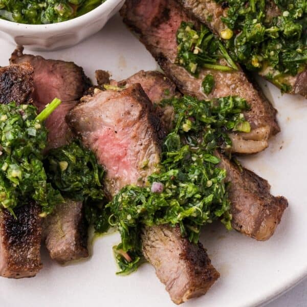 Sliced grilled steak with chimichurri, topped with vibrant green sauce and served on a white plate.