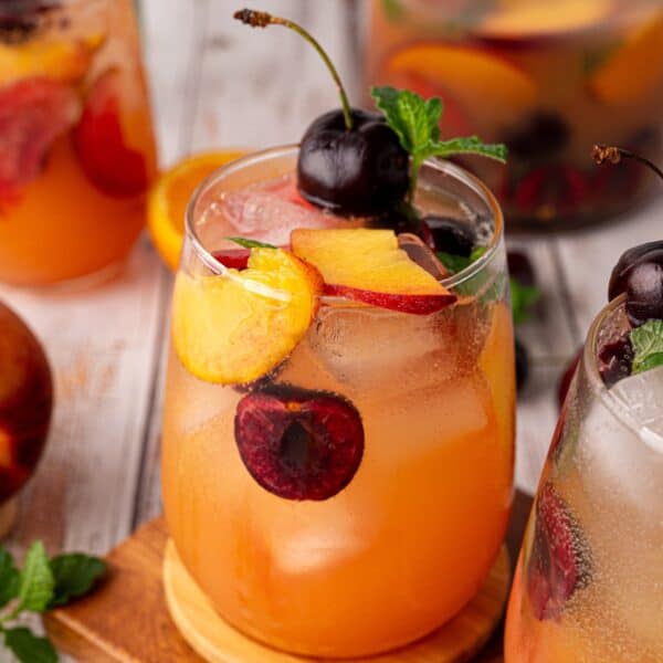 A glass of iced stone fruit mocktail featuring peach and cherry, garnished with fresh fruit slices and mint, sits on a wooden surface.