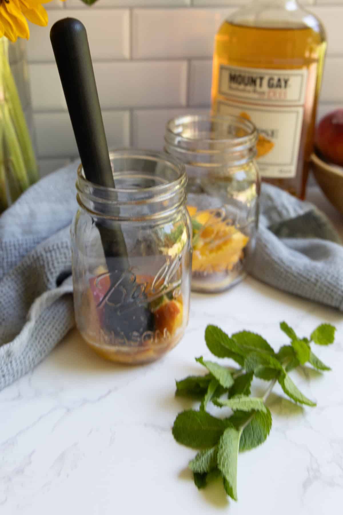 Two mason jars with muddled fruit for a bourbon peach smash cocktail sit on a countertop, a muddler nearby. A bottle of Mount Gay rum is in the background, with mint leaves and a gray cloth completing the scene.