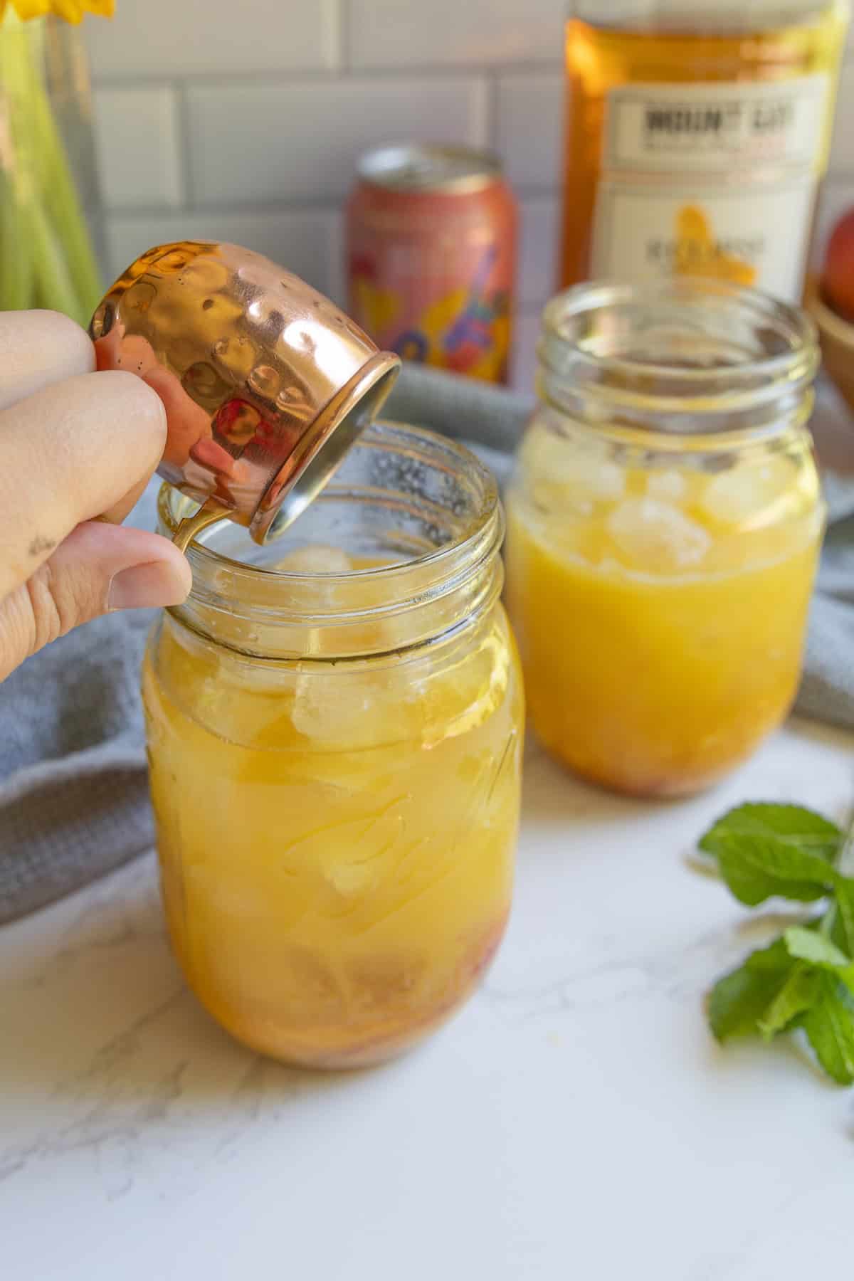 A hand pours liquid from a small copper cup into a mason jar filled with ice and an orange bourbon peach smash cocktail; another similar jar sits nearby on the marble counter.