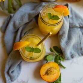 Two mason jars of bourbon peach smash cocktail with ice, garnished with mint and peach slices, placed on a light cloth, with a halved peach and mint sprigs nearby.