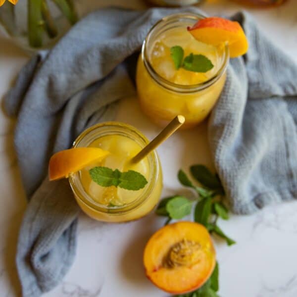 Two mason jars of bourbon peach smash cocktail with ice, garnished with mint and peach slices, placed on a light cloth, with a halved peach and mint sprigs nearby.