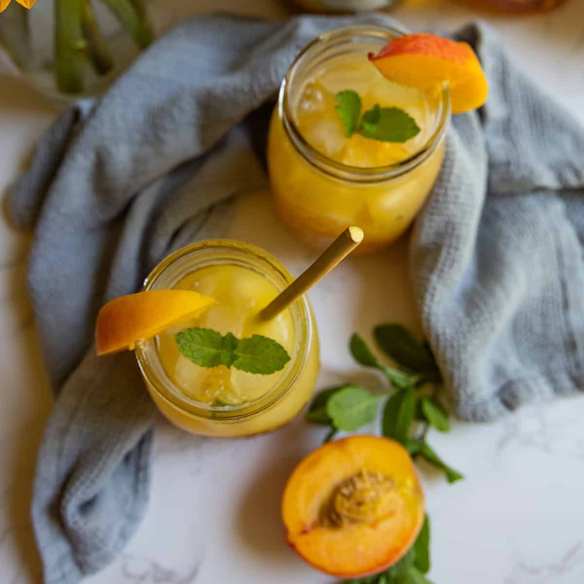 Two mason jars of bourbon peach smash cocktail with ice, garnished with mint and peach slices, placed on a light cloth, with a halved peach and mint sprigs nearby.
