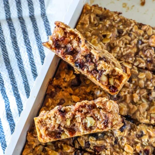 Close-up of sourdough granola bars with oats, nuts, and visible dried fruit, stacked in a baking dish next to a striped towel.