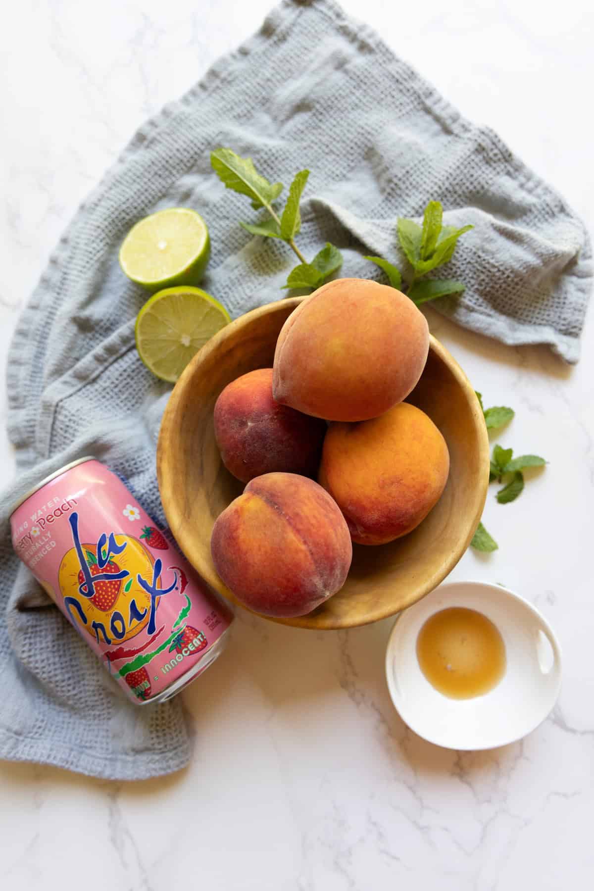 A wooden bowl with peaches, a can of La Croix, fresh mint, lime halves, and a small dish of honey are arranged on a cloth—perfect ingredients for crafting a refreshing peach La Croix mocktail.