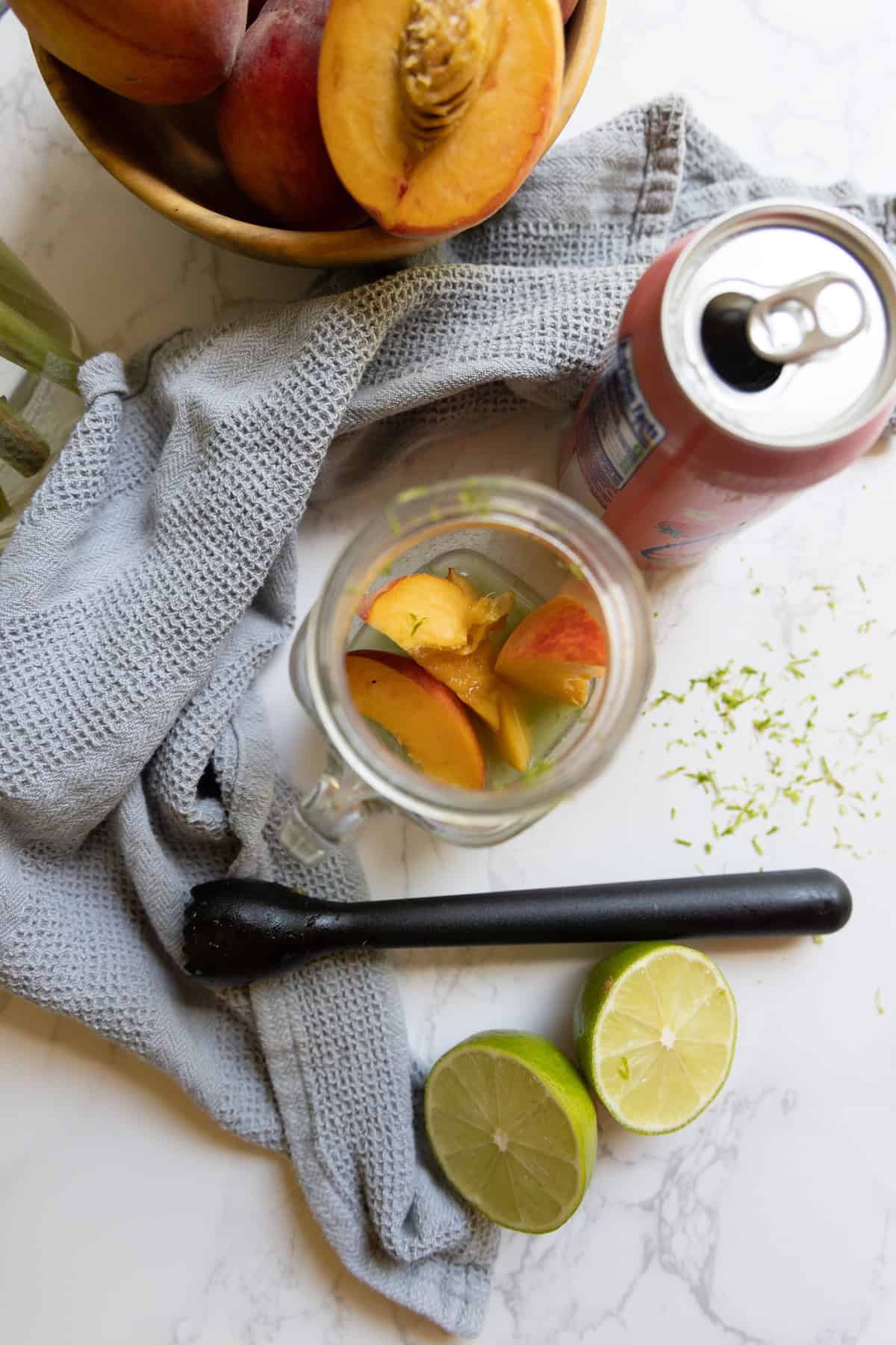 A mason jar with peach slices, a halved lime, a peach, a can of Peach La Croix mocktail, a black muddler, a gray cloth, and a bowl of peaches rest on a marble surface.