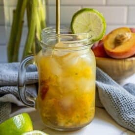 A mason jar filled with a refreshing peach La Croix mocktail, garnished with a lime wheel and served with a straw; peaches and limes are visible in the background.