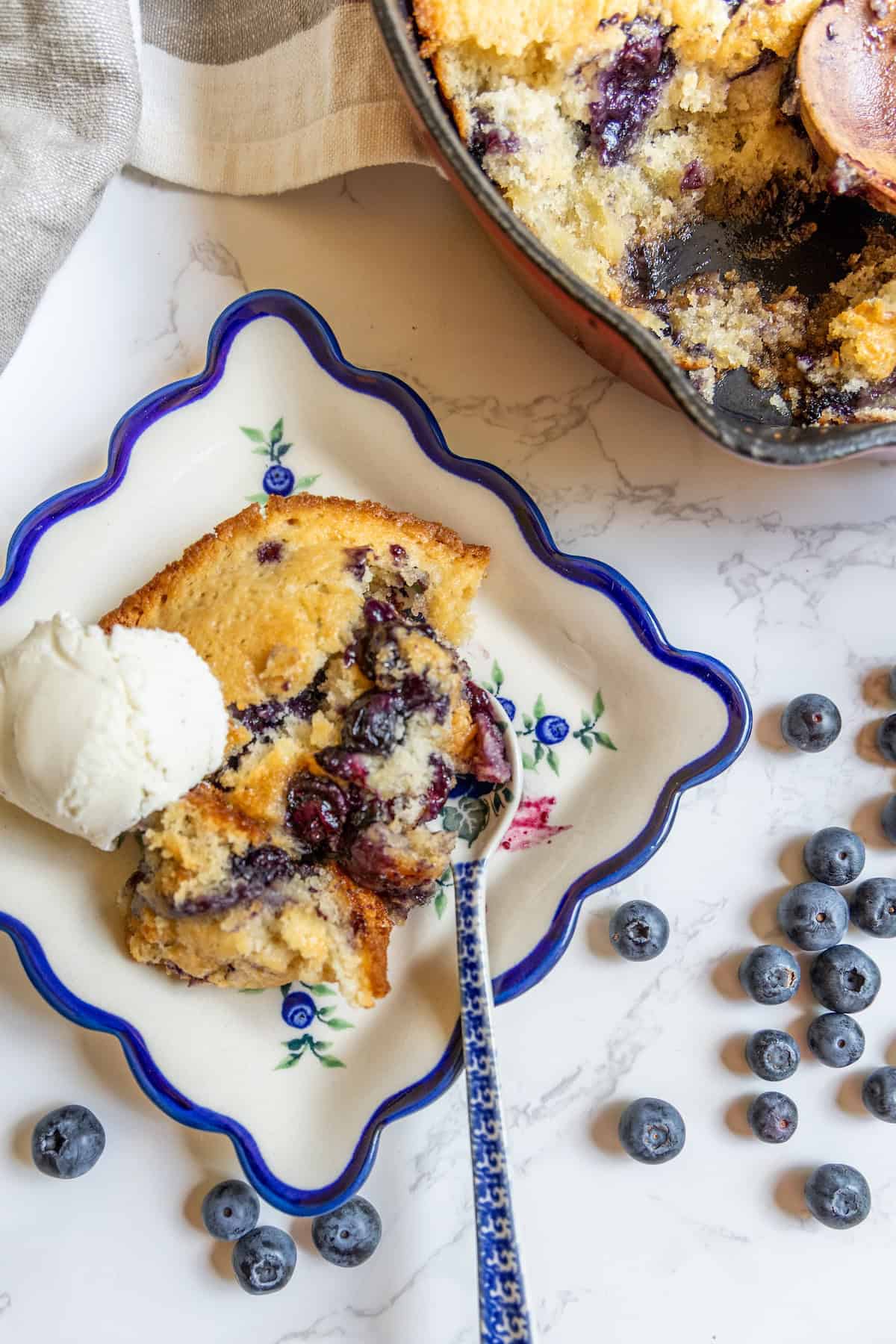 A slice of sourdough blueberry cobbler with a scoop of vanilla ice cream on a decorative plate, fresh blueberries scattered nearby.