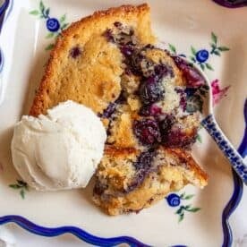 A slice of sourdough blueberry cobbler with a scoop of vanilla ice cream sits on a decorative plate, with a spoon resting nearby.