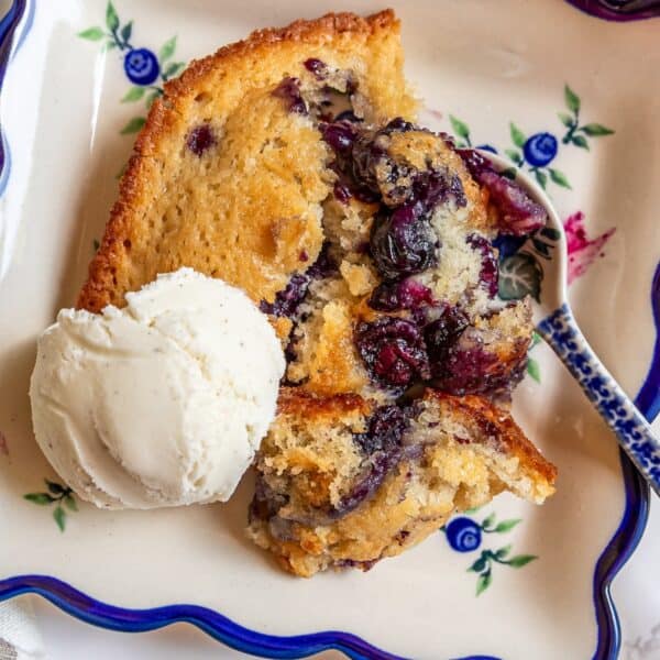 A slice of sourdough blueberry cobbler with a scoop of vanilla ice cream sits on a decorative plate, with a spoon resting nearby.