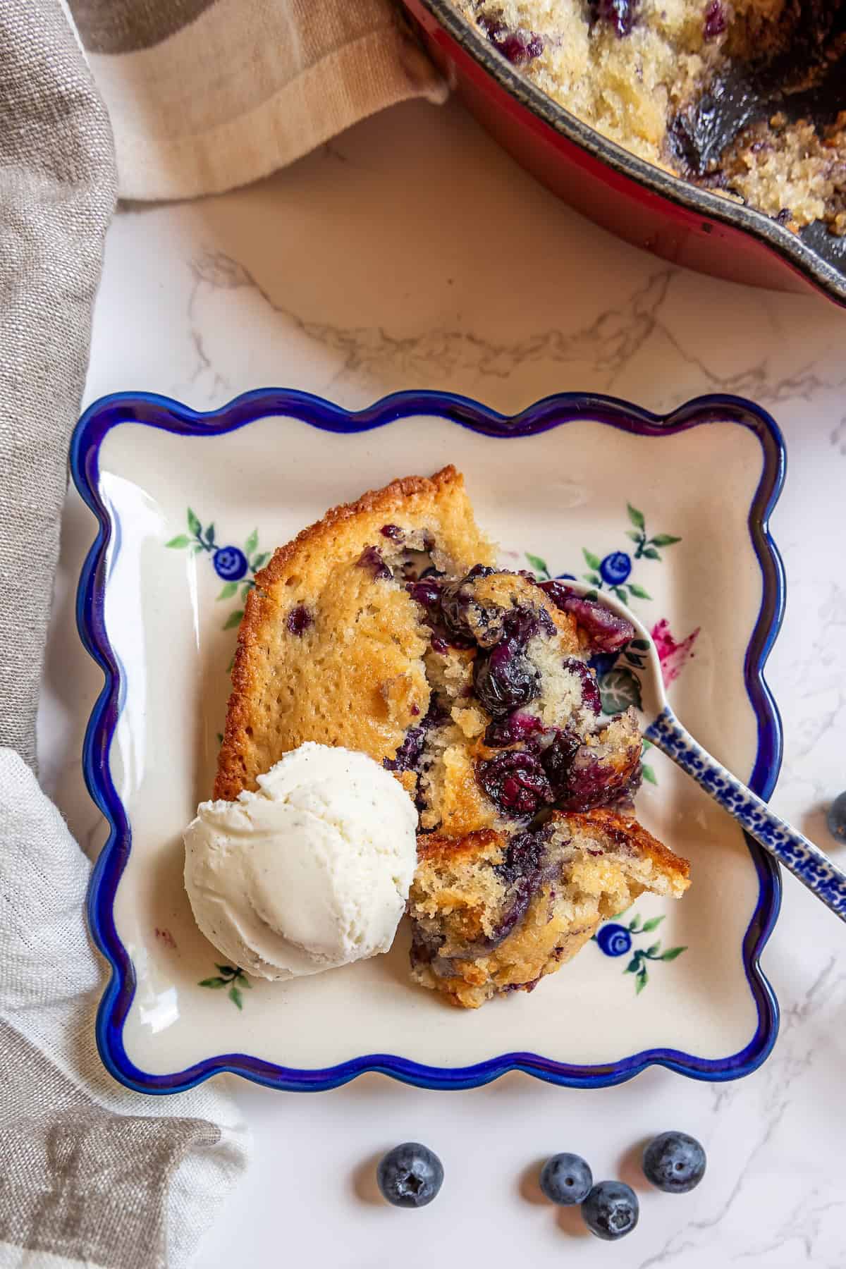 Sour-Sweet Sourdough Blueberry Cobbler 2 A slice of sourdough blueberry cobbler with a scoop of vanilla ice cream on a decorative plate, with a few blueberries nearby.
