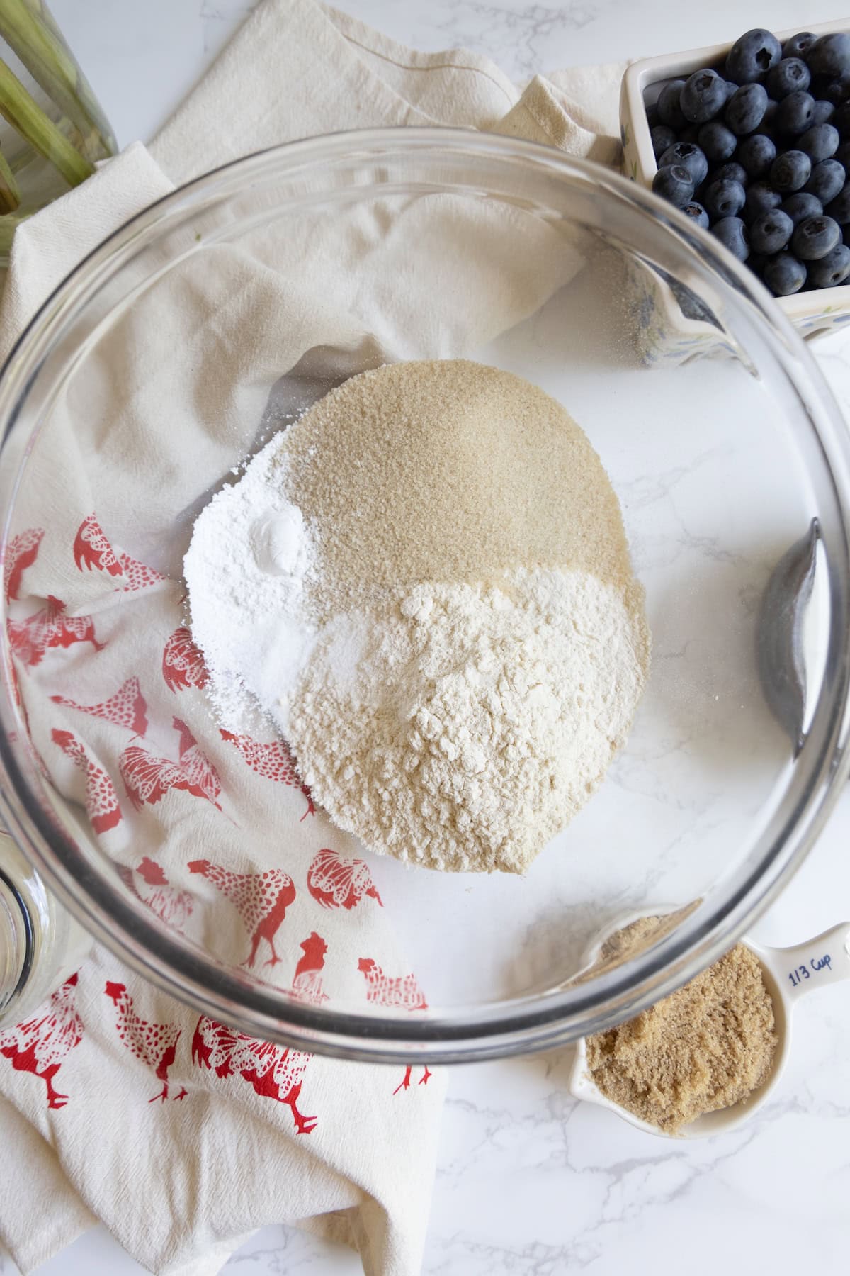A glass bowl containing flour, sugar, and baking powder sits on a counter near a container of blueberries and a dish of brown sugar—ingredients ready for a homemade sourdough blueberry cobbler.
