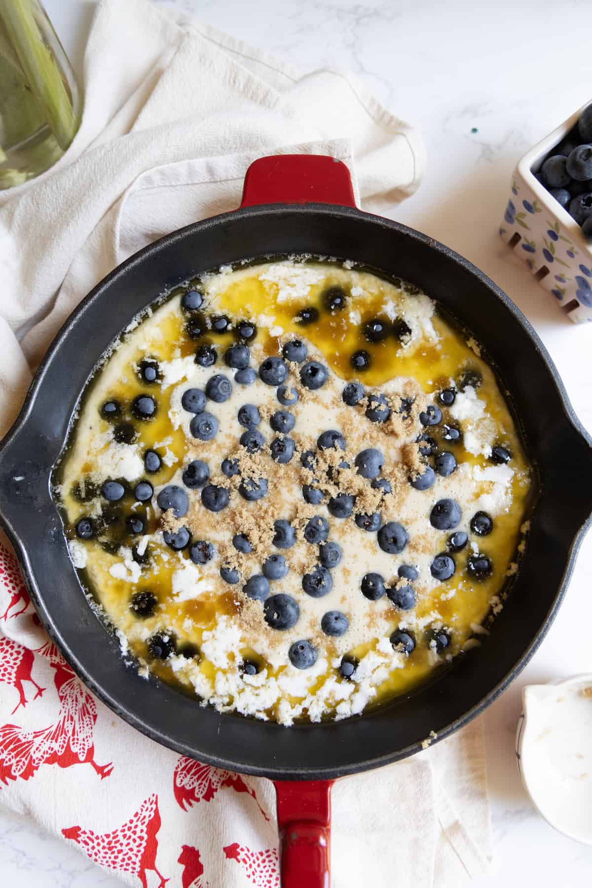 A cast iron skillet filled with unbaked sourdough blueberry cobbler—batter, melted butter, blueberries, and brown sugar—rests on a white countertop with a towel and a container of blueberries nearby.