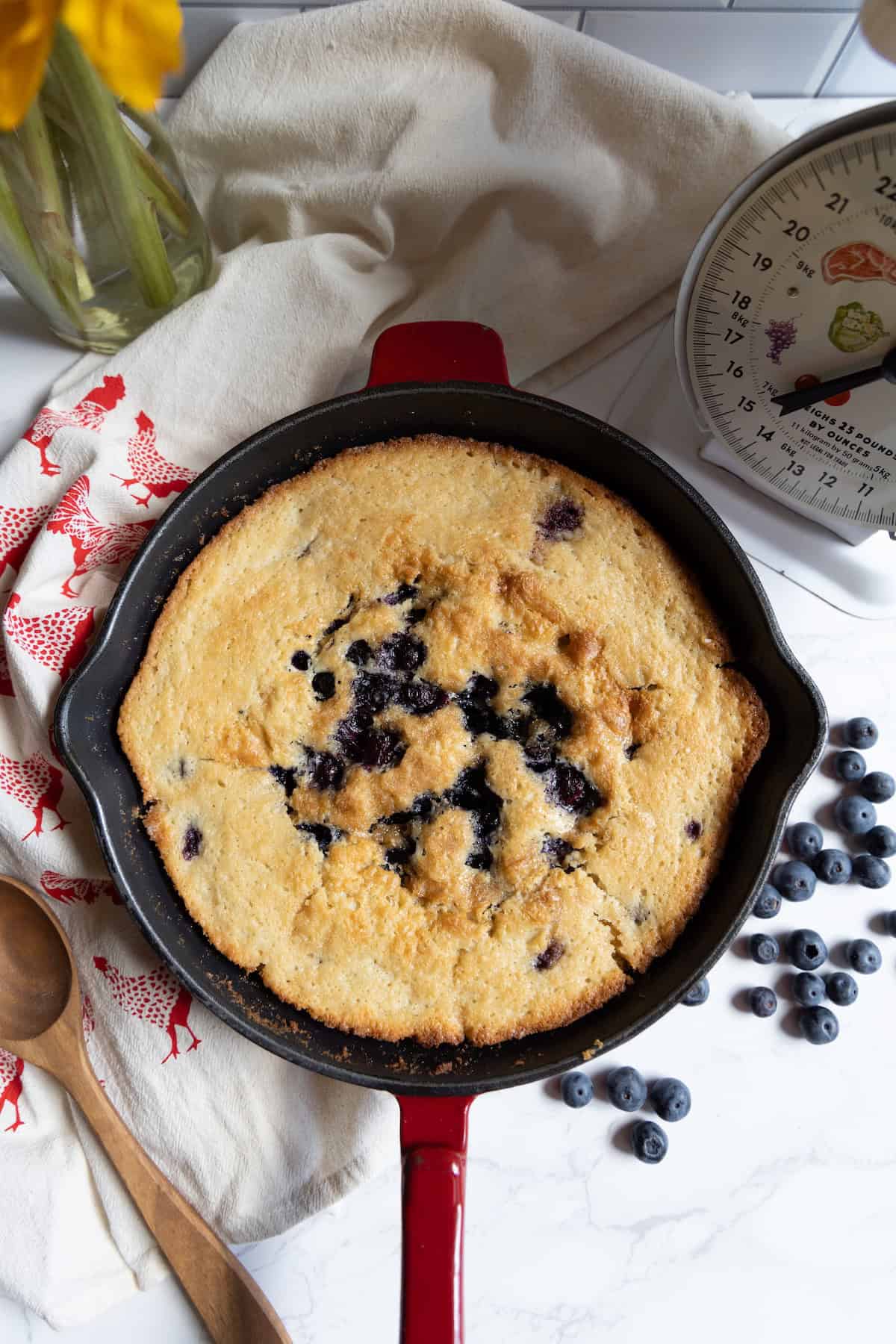 A baked sourdough blueberry cobbler skillet cornbread sits in a red cast iron pan on a marble countertop, with fresh blueberries, a kitchen scale, wooden spoons, and a napkin nearby.