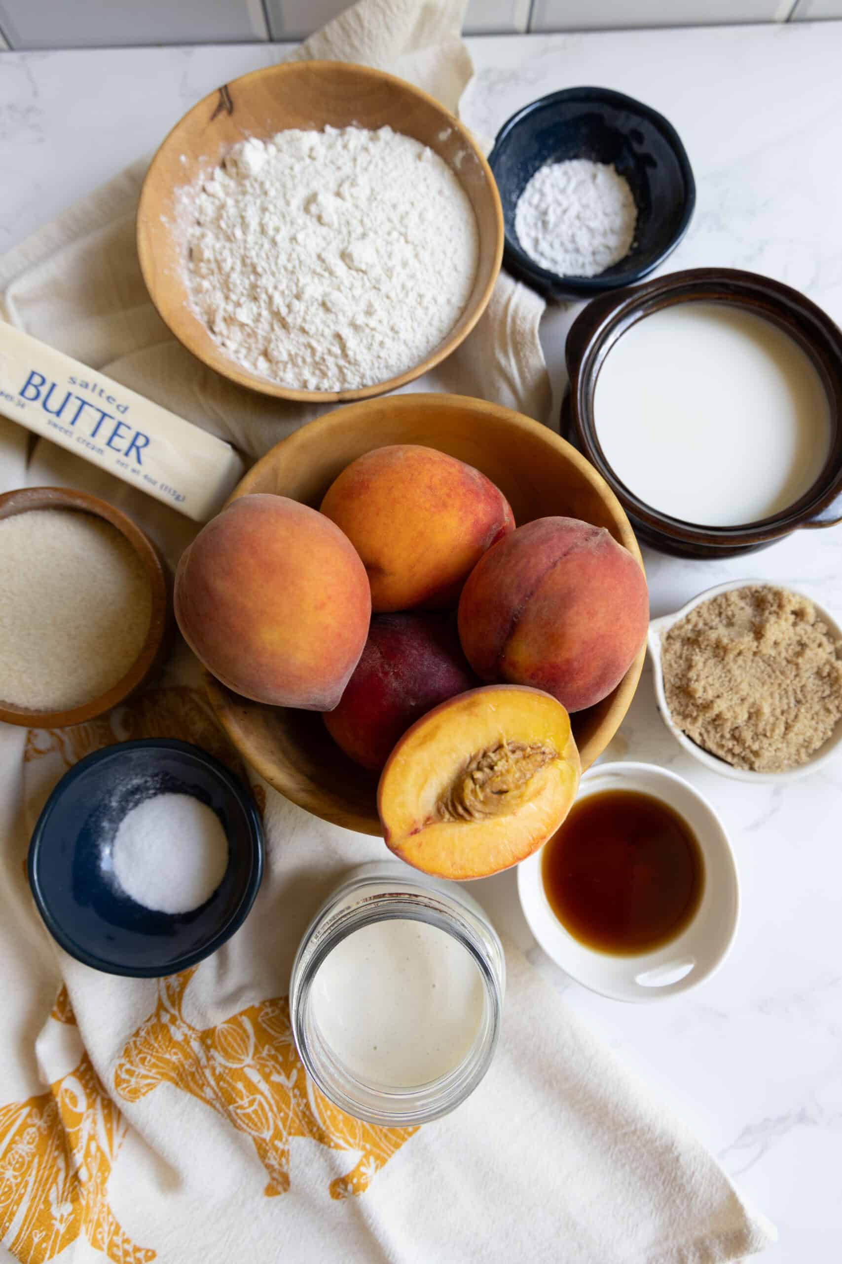 A flat lay of baking ingredients for sourdough peach cobbler includes fresh peaches, flour, sugar, brown sugar, butter, milk, vanilla extract, baking powder, and salt on a kitchen towel.