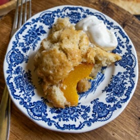 A slice of sourdough peach cobbler with a dollop of whipped cream sits on a blue and white floral-patterned plate, next to a fork on a wooden surface.