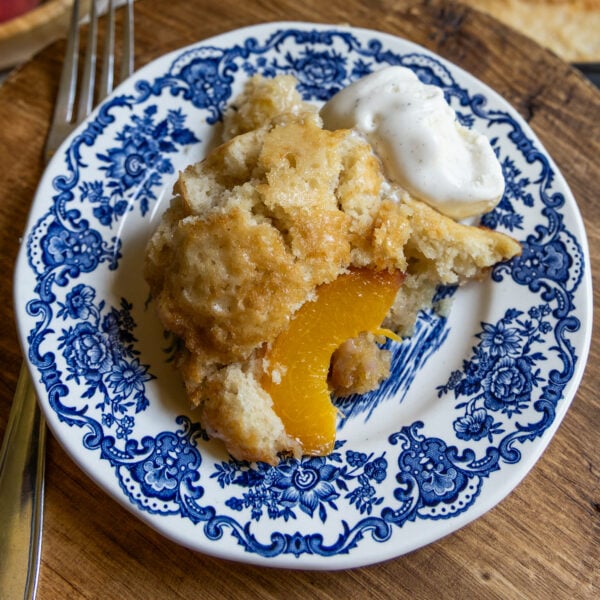 A slice of sourdough peach cobbler with a dollop of whipped cream sits on a blue and white floral-patterned plate, next to a fork on a wooden surface.