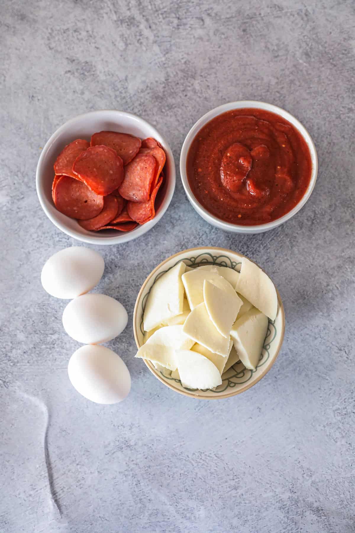 Three eggs, a bowl of sliced pepperoni, a bowl of pizza sauce, and mozzarella cheese are arranged on a grey surface—everything you need for a delicious keto breakfast pizza.