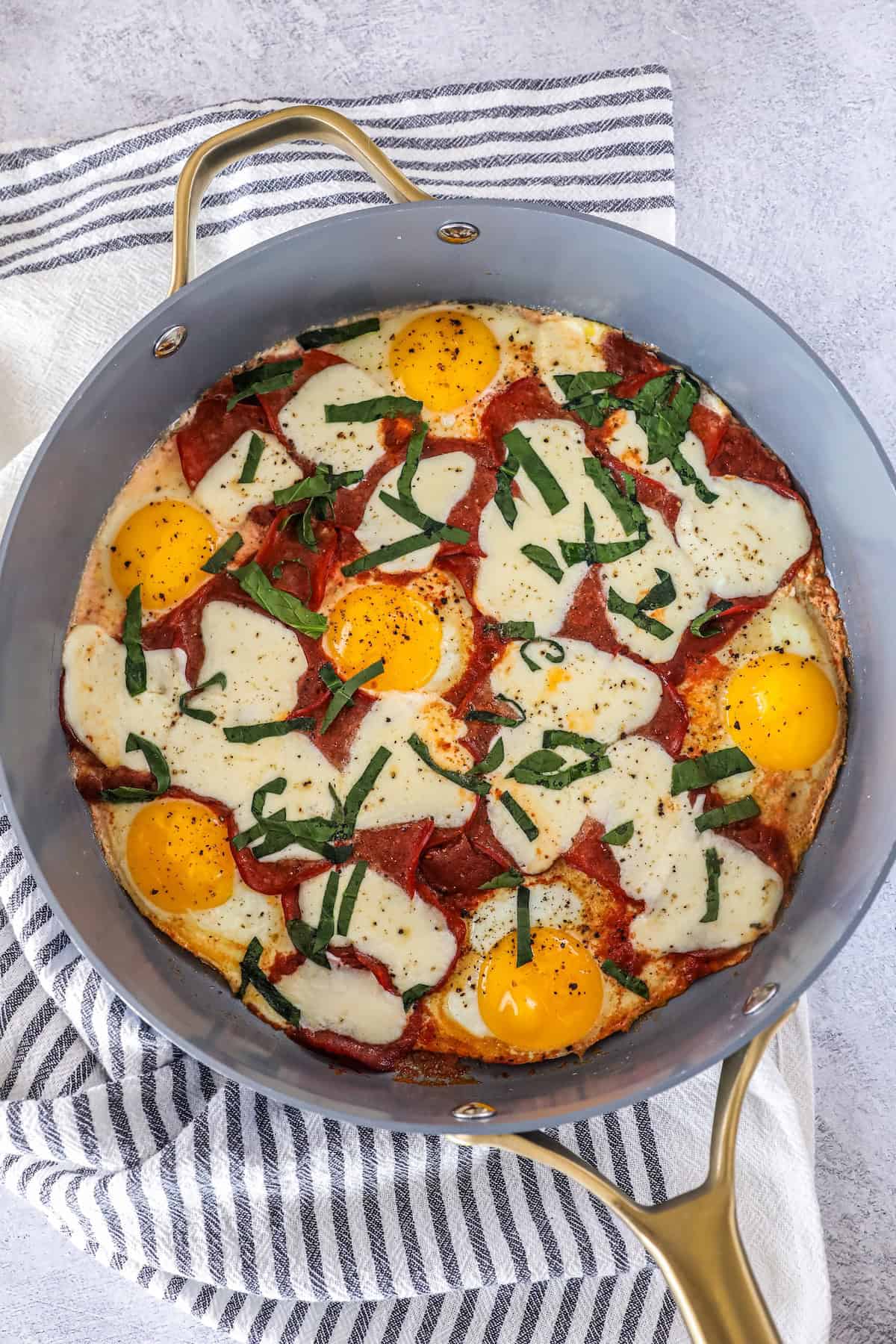 A cooked keto breakfast pizza in a skillet with sunny-side-up eggs, melted cheese, pepperoni slices, and chopped herbs, placed on a striped towel.
