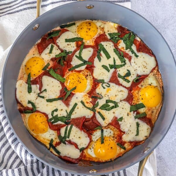 A skillet containing eggs, melted cheese, pepperoni slices, and chopped basil, with ground pepper sprinkled on top—this keto breakfast pizza is a low-carb twist on a classic favorite.