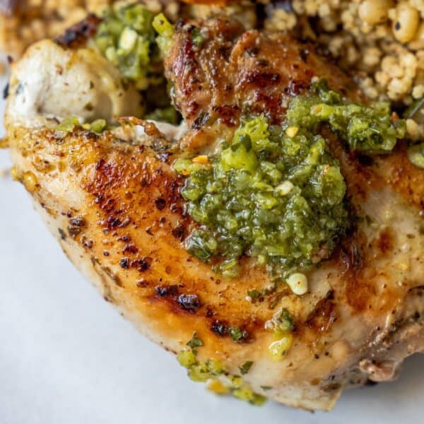 Close-up of juicy chicken thighs topped with vibrant zhoug sauce, served alongside a portion of grain salad.