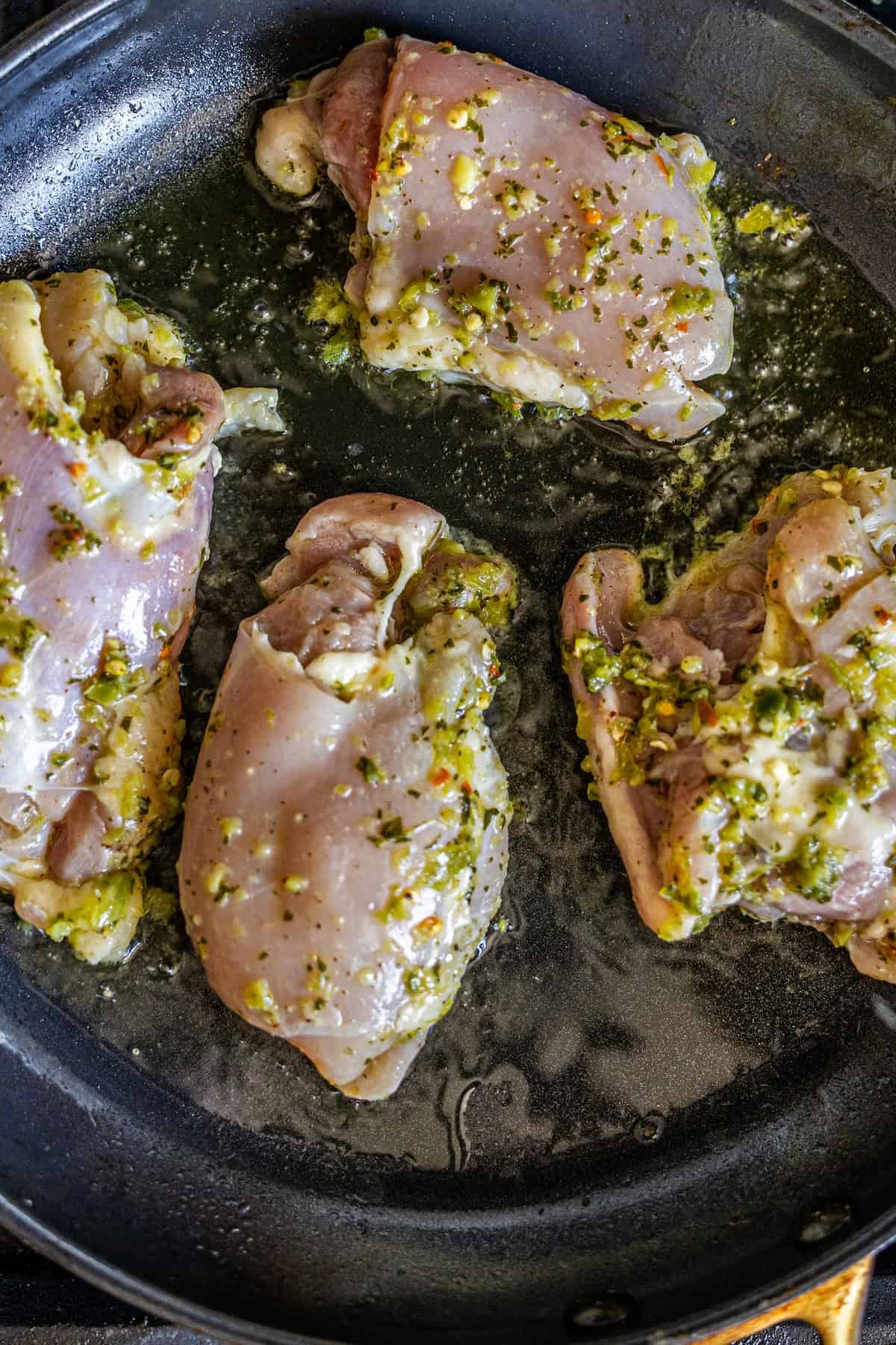 Raw, marinated Zhoug Chicken thighs sizzling in oil in a black skillet.