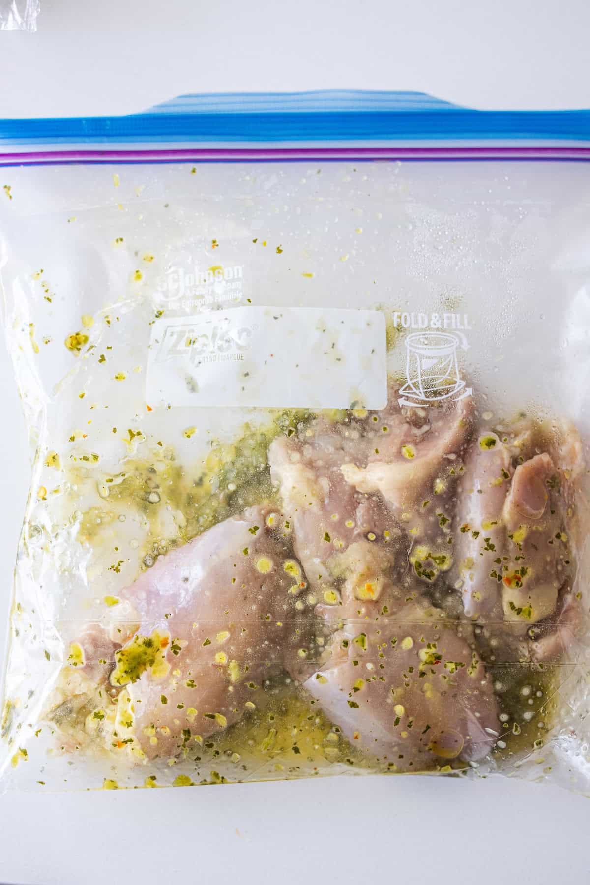 Zhoug Chicken pieces marinating in a sealed plastic zip-top bag with visible herbs and seasoning.