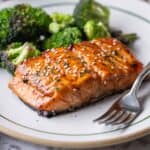 A plate with Japanese BBQ Salmon, glazed and topped with sesame seeds, served alongside roasted broccoli, with a fork placed on the side.