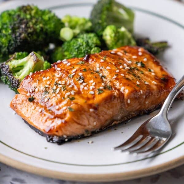 A plate with Japanese BBQ Salmon, glazed and topped with sesame seeds, served alongside roasted broccoli, with a fork placed on the side.