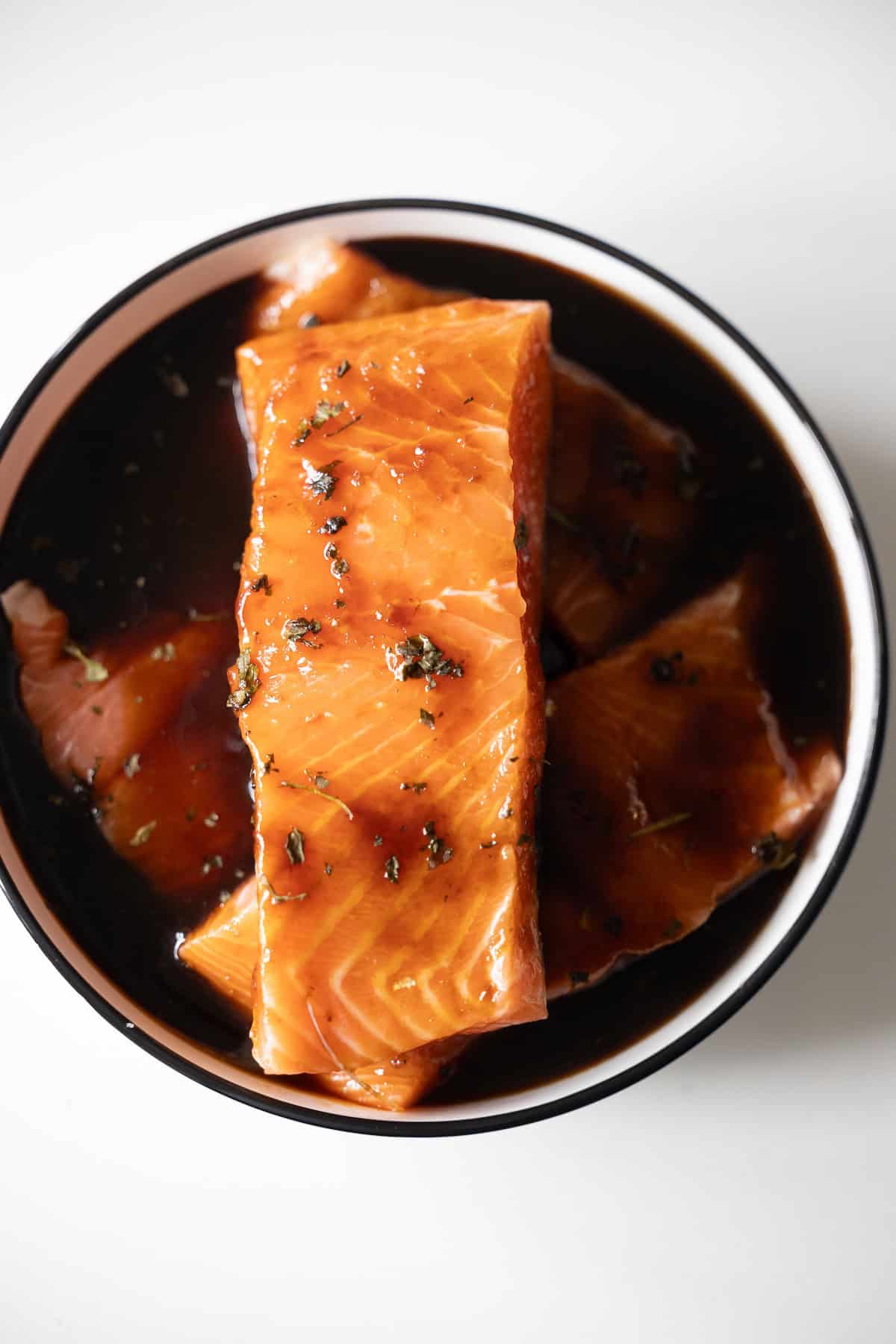 Japanese BBQ Salmon fillets marinate in a dark, flavorful sauce with herbs, arranged in a round bowl on a white surface.